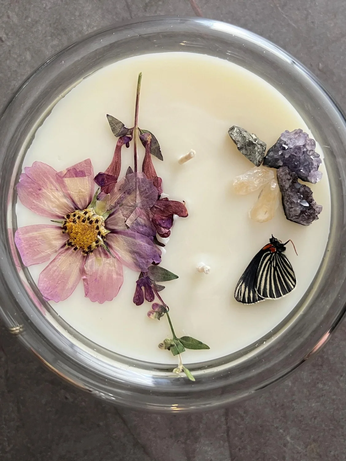 Vista superior de una vela Moon Cristal: flor de cosmos rosa prensada, cristales de amatista y una mariposa sobre la cera